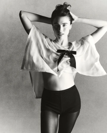 Black-and-white portrait of a fair-skinned woman wearing a sheer white top with a black bow, shorts, and stockings, with hands in her hair.
