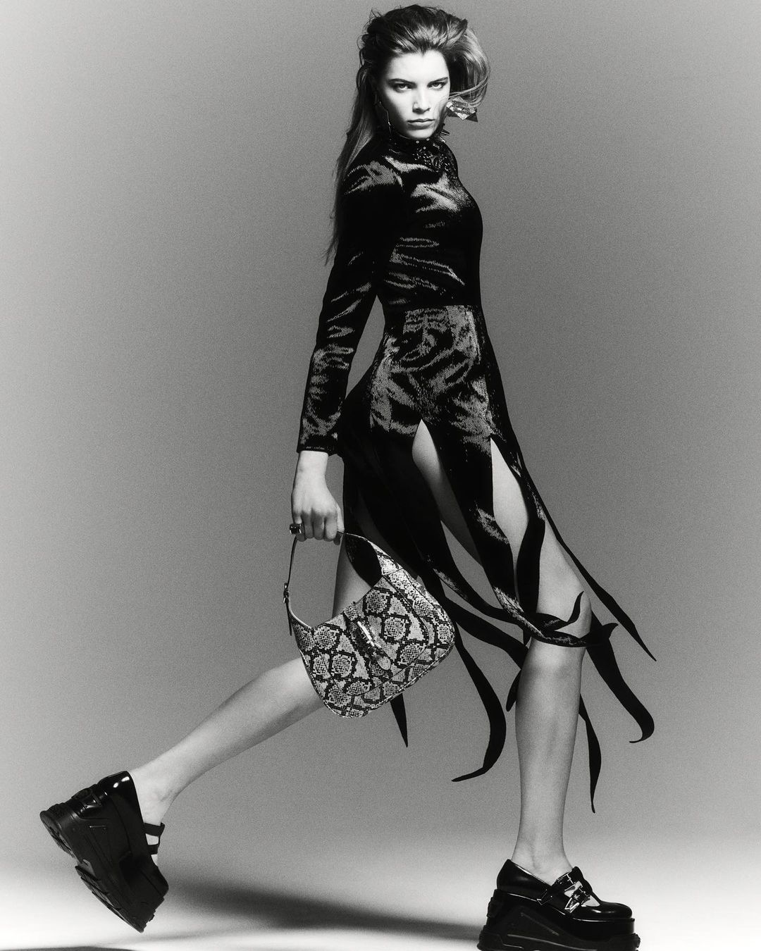 A confident woman strides forward in a striking black dress with cutouts, chunky platform shoes, and a textured bag against a grey backdrop.