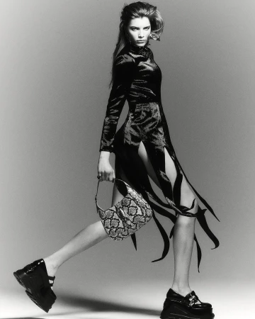 A confident woman strides forward in a striking black dress with cutouts, chunky platform shoes, and a textured bag against a grey backdrop.