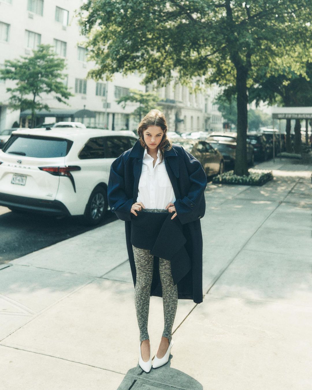 A stylish woman in a long blue coat, white shirt, and patterned stirrup leggings poses on a city sidewalk with buildings in the background.