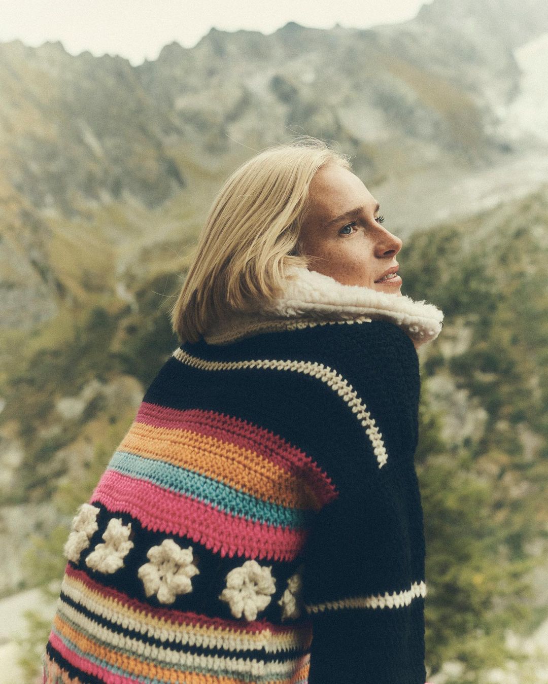 Woman in colorful knit sweater with mountainous landscape background, outdoors. Female fashion portrait in nature setting, back view.