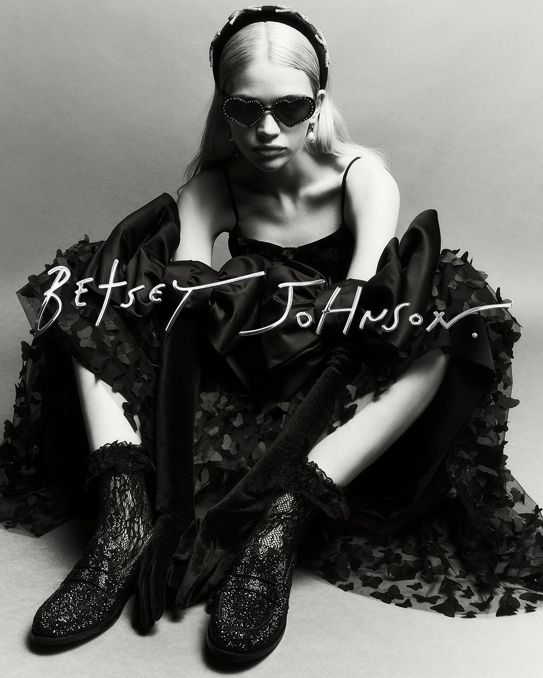 Young woman in a black dress, heart-shaped sunglasses, and platform shoes poses in a studio against a grey backdrop. Fashion shot.