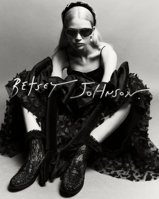 Young woman in a black dress, heart-shaped sunglasses, and platform shoes poses in a studio against a grey backdrop. Fashion shot.