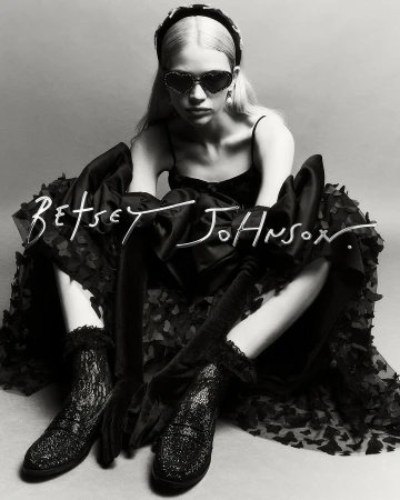 Young woman in a black dress, heart-shaped sunglasses, and platform shoes poses in a studio against a grey backdrop. Fashion shot.