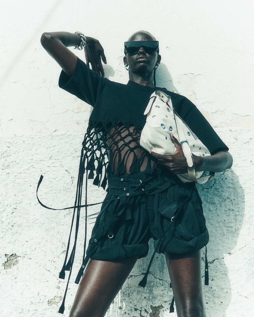 A slender Black female model poses in a black crop top, shorts, sunglasses, earrings, and bracelet holding a white bag near a white wall.
