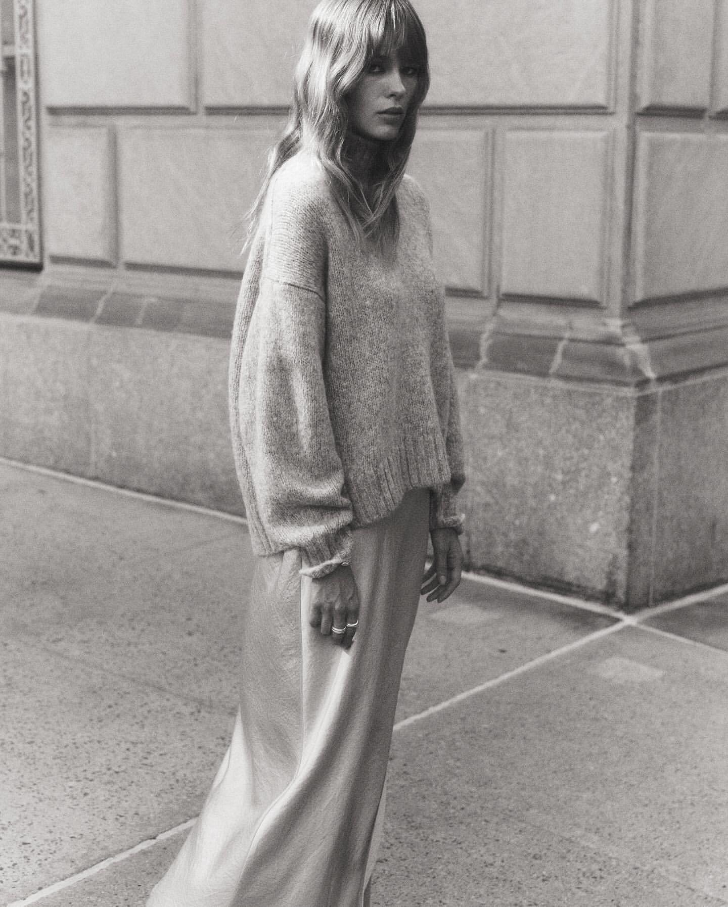 Monochrome portrait of a woman with long hair wearing a sweater and silk skirt, standing in front of a building on a city street.