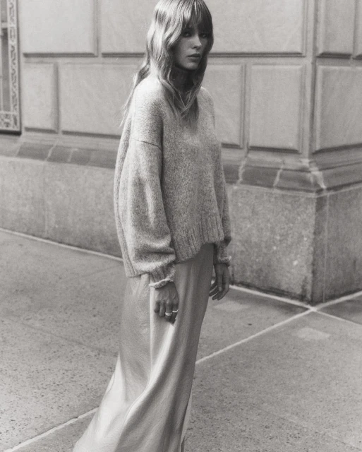 Monochrome portrait of a woman with long hair wearing a sweater and silk skirt, standing in front of a building on a city street.