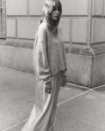 Monochrome portrait of a woman with long hair wearing a sweater and silk skirt, standing in front of a building on a city street.