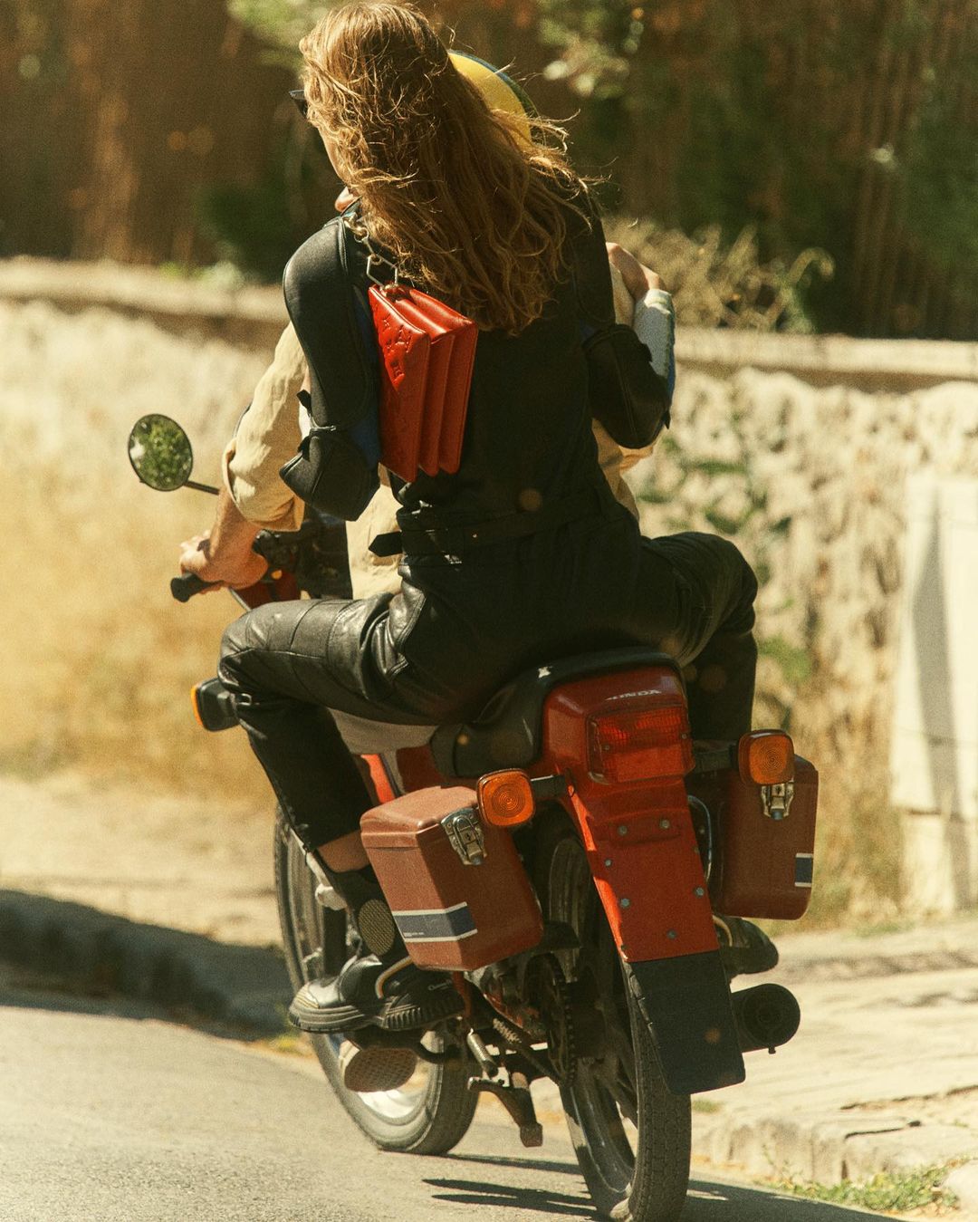 Two people ride a vintage red motorcycle on a sunny road with a red leather bag and side cases, capturing a retro and adventurous vibe.