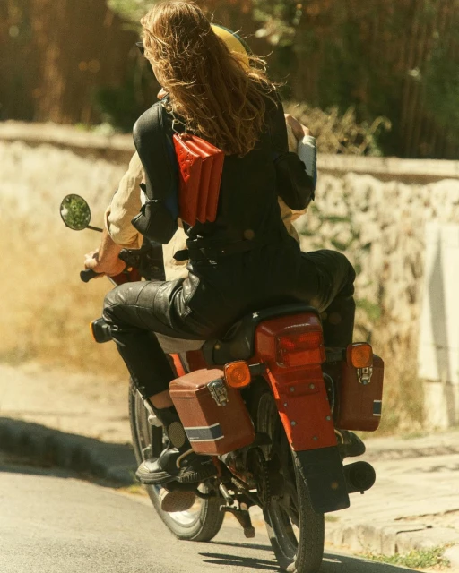 Two people ride a vintage red motorcycle on a sunny road with a red leather bag and side cases, capturing a retro and adventurous vibe.