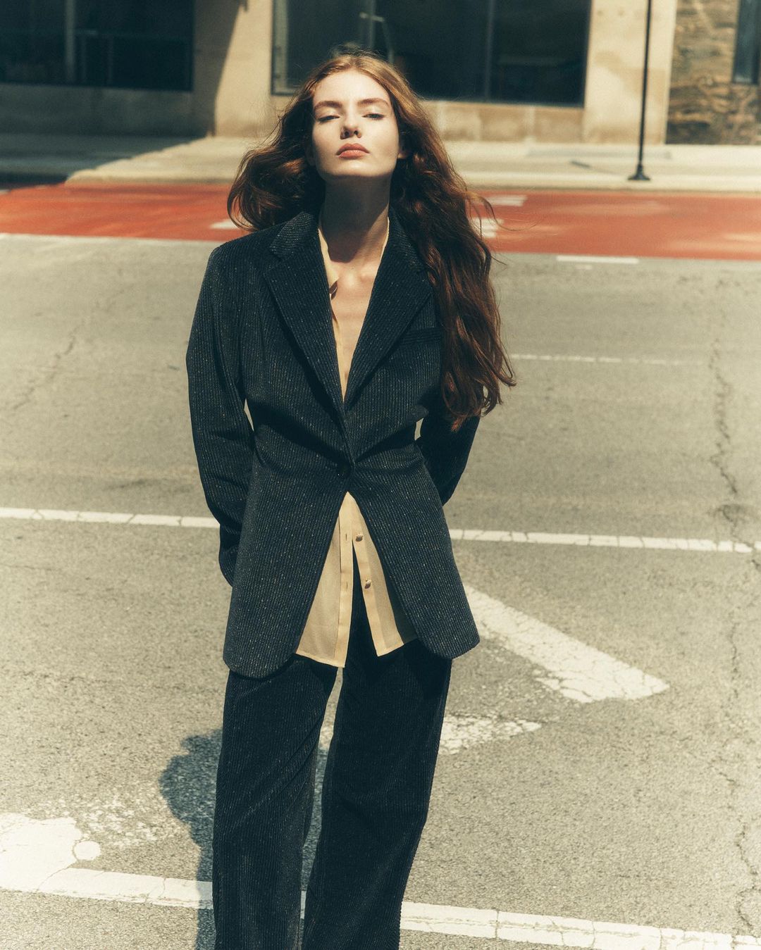 Woman in a dark blazer and pants stands on a city street with an orange crosswalk and tall buildings in the background, fashion shot.
