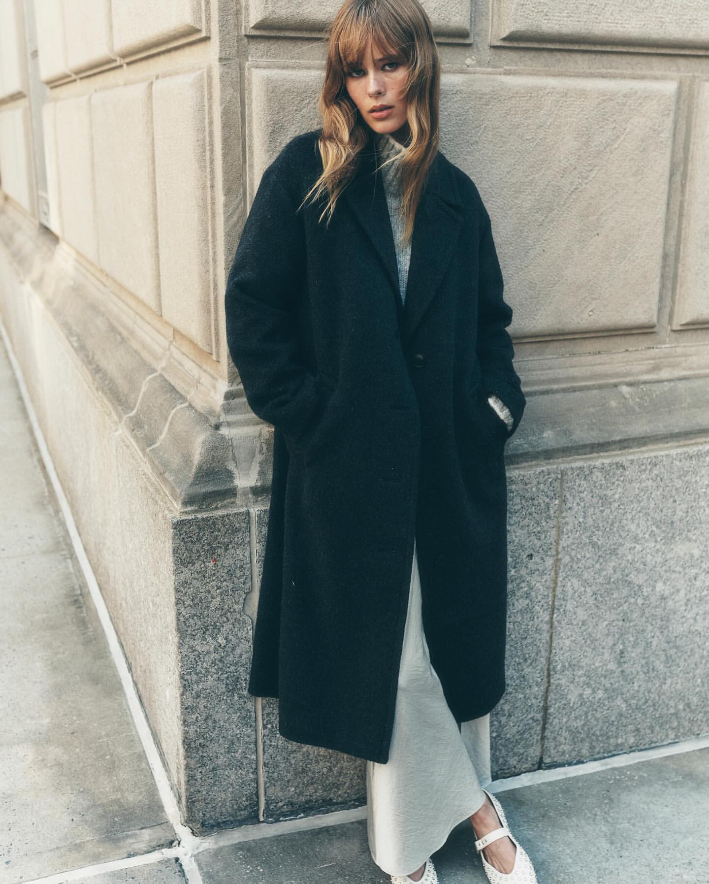 A stylish adult female poses outdoors against a building in a full-length black overcoat with cream-colored knit pants and white shoes.