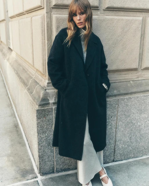 A stylish adult female poses outdoors against a building in a full-length black overcoat with cream-colored knit pants and white shoes.