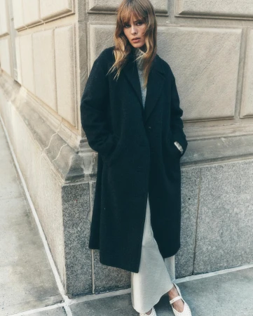 A stylish adult female poses outdoors against a building in a full-length black overcoat with cream-colored knit pants and white shoes.