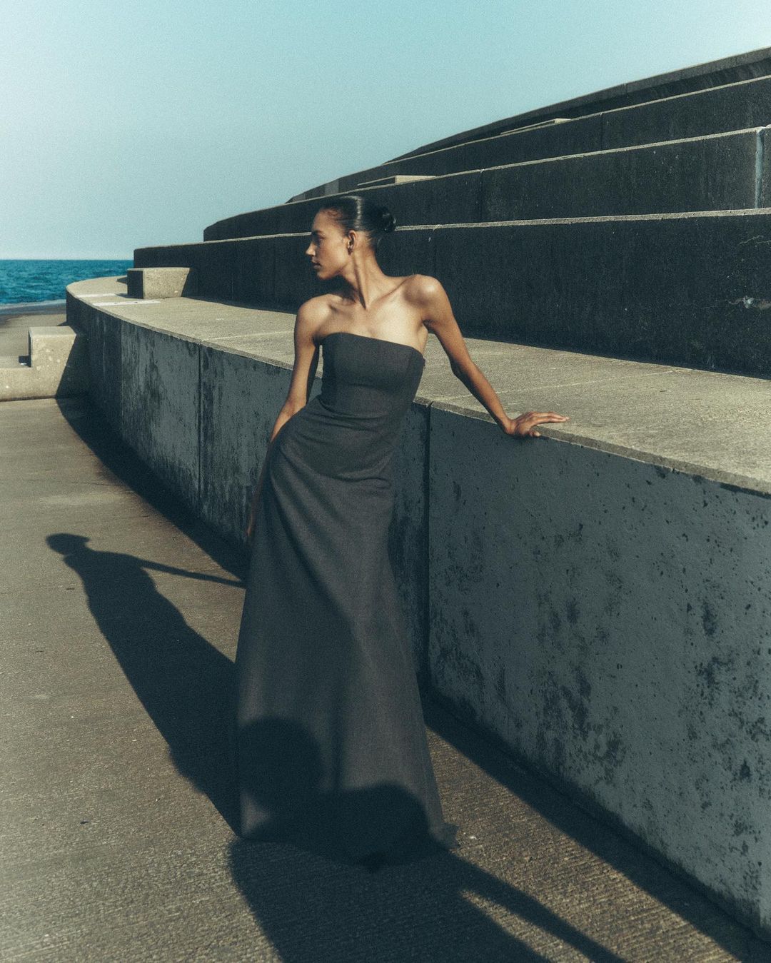 Elegant woman in strapless gown poses on an outdoor concrete structure with ocean view, side lit by sunlight casting long shadow.