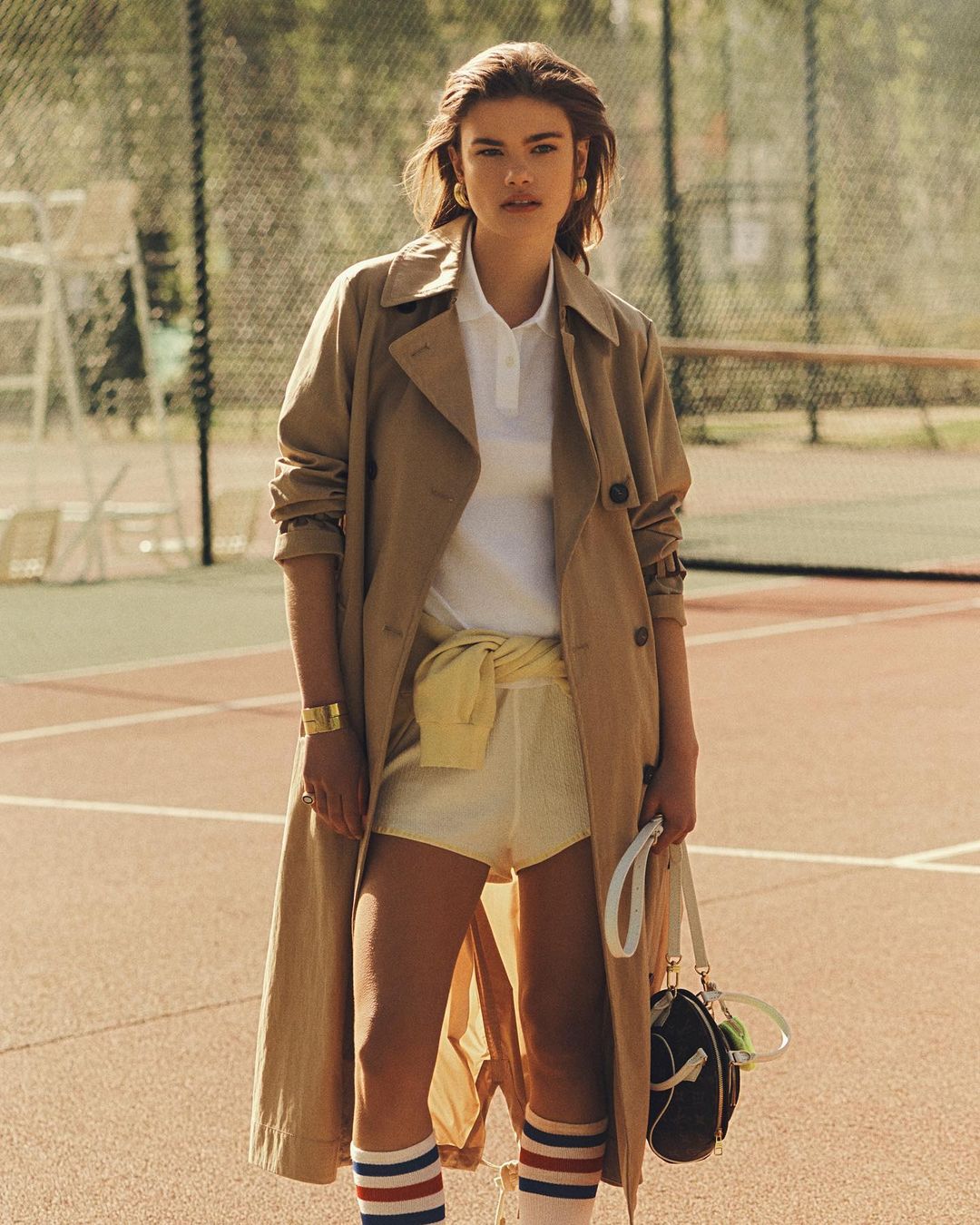 Woman in trench coat poses on tennis court. She wears a white polo, shorts, and tube socks with a sports bag and a tennis net behind.