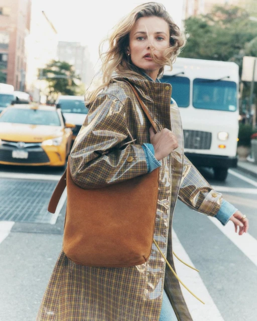 Woman in a transparent plaid trench coat and suede tote bag crosses a city street with yellow taxi cabs and white truck in background.
