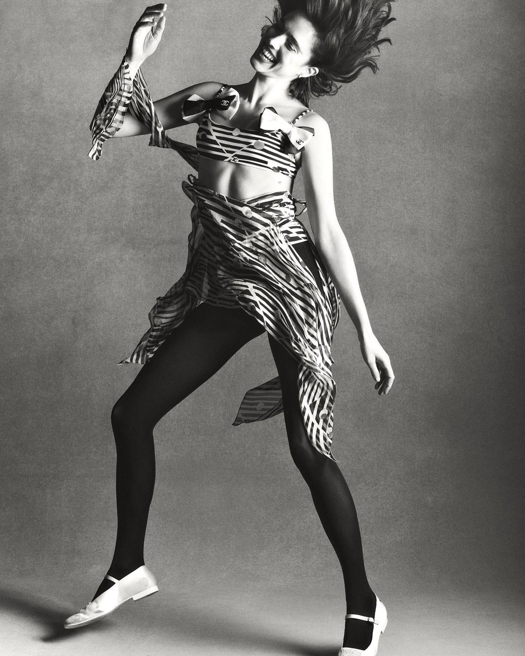 Monochrome studio shot of a woman in a striped two-piece outfit, tights, and Mary Jane shoes, jumping against a grey backdrop.