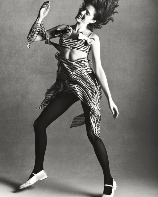 Monochrome studio shot of a woman in a striped two-piece outfit, tights, and Mary Jane shoes, jumping against a grey backdrop.