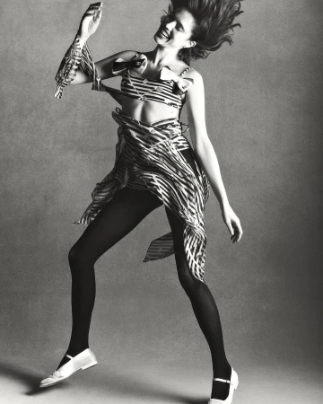 Monochrome studio shot of a woman in a striped two-piece outfit, tights, and Mary Jane shoes, jumping against a grey backdrop.