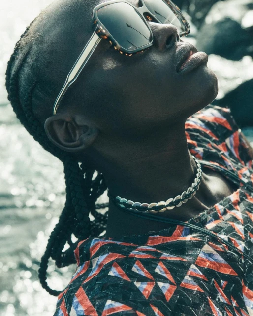 Woman in sunglasses wearing a patterned top and silver necklace, looking up at the sky, with water and rocks in the background.