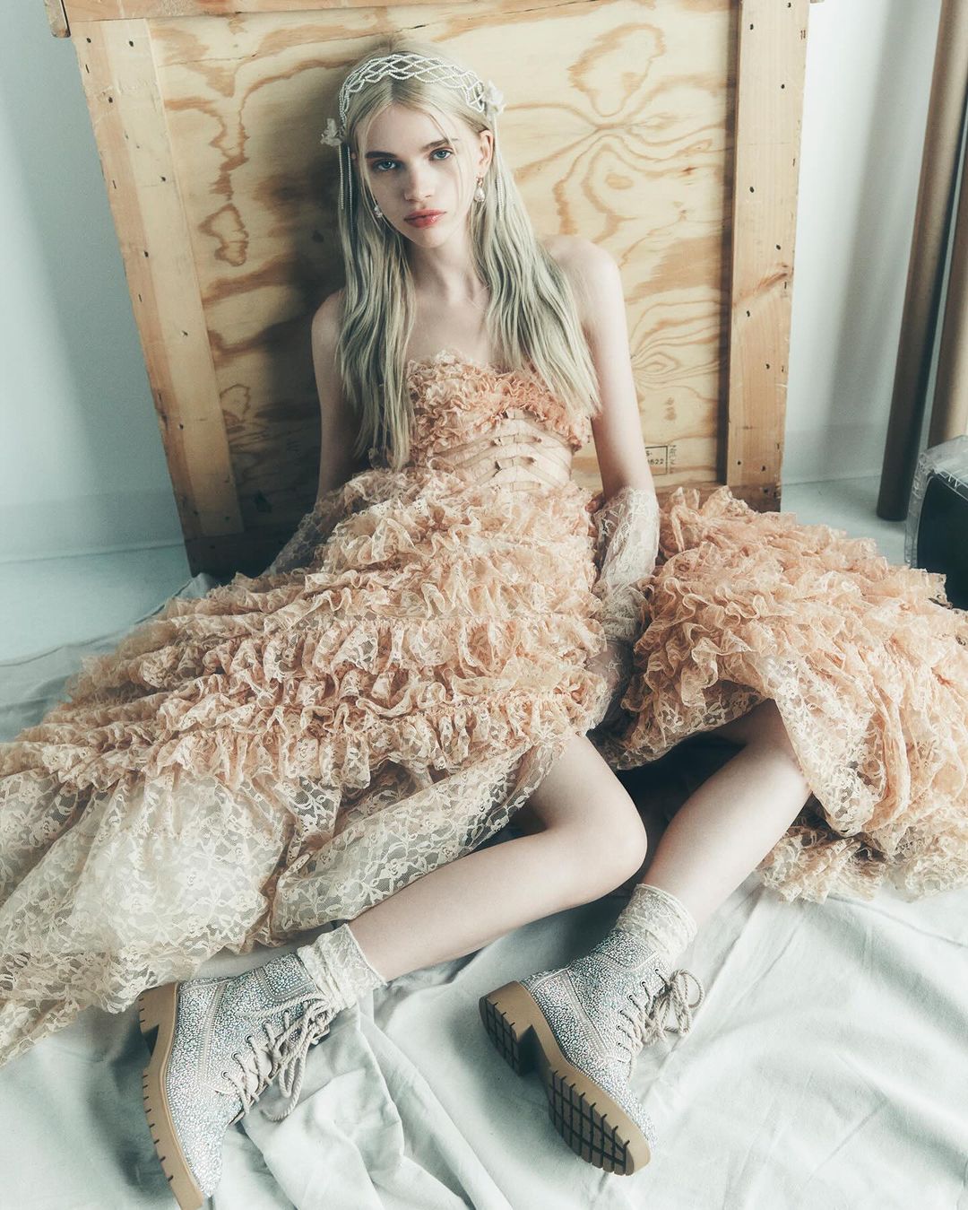 Young woman poses in a ruffled peach-colored dress and embellished boots, seated against a wooden backdrop in soft, diffused lighting.