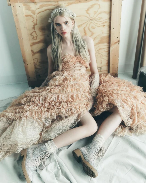 Young woman poses in a ruffled peach-colored dress and embellished boots, seated against a wooden backdrop in soft, diffused lighting.