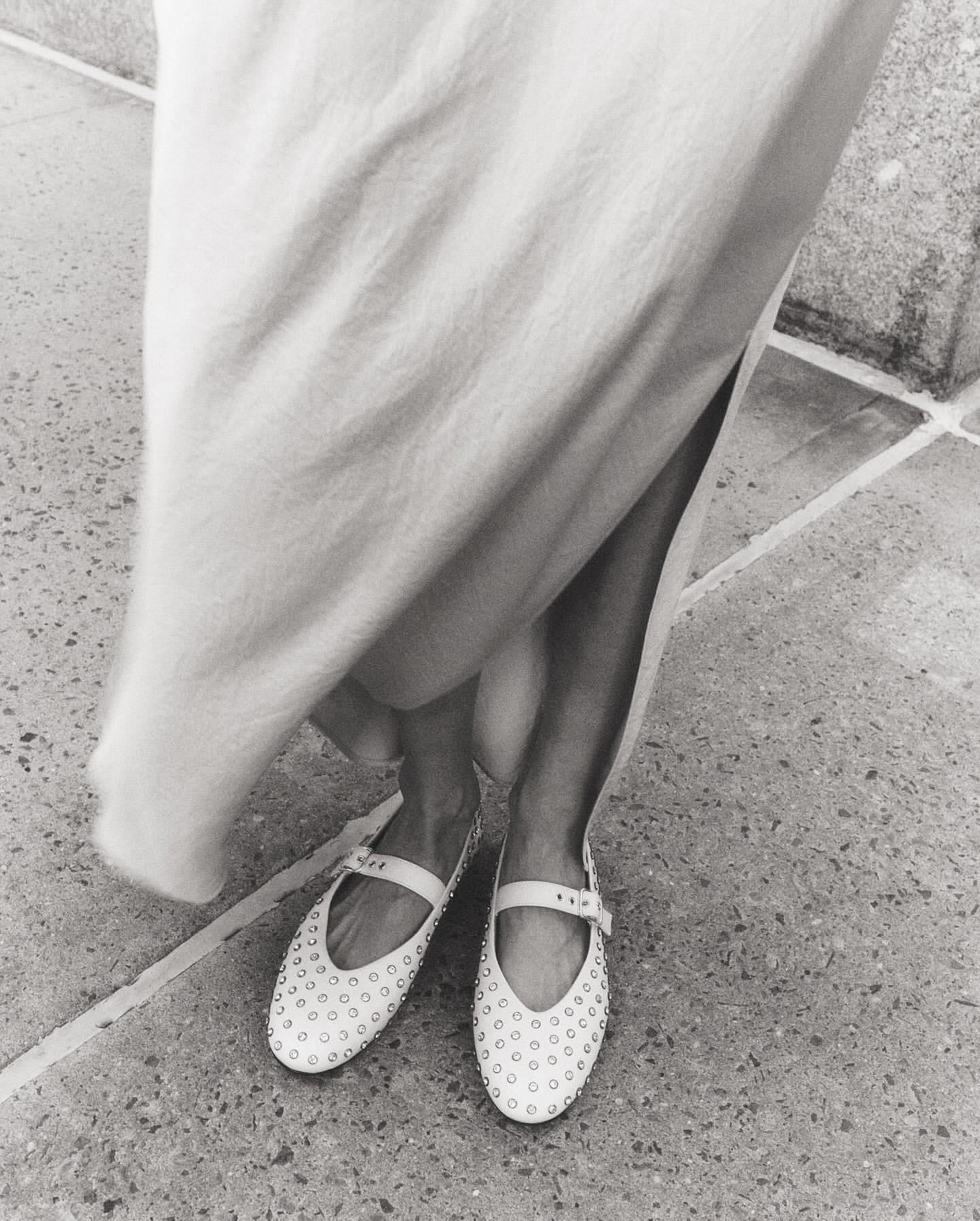 Monochrome shot of woman's legs and feet wearing studded flats with ankle straps and a light dress on a tiled surface.