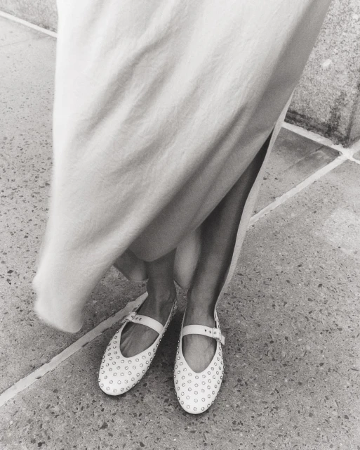 Monochrome shot of woman's legs and feet wearing studded flats with ankle straps and a light dress on a tiled surface.