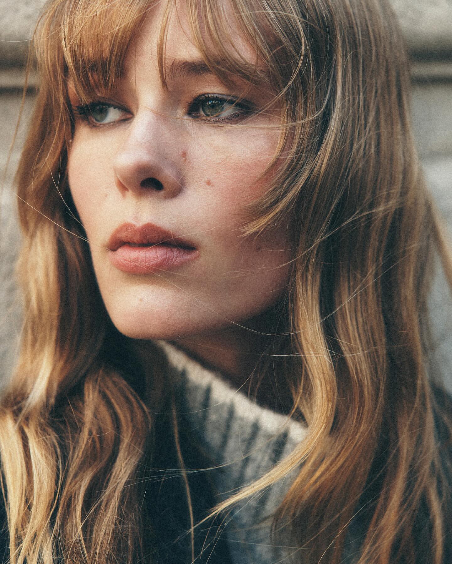 Close-up portrait of a young, fair-skinned woman with wavy blonde hair and neutral expression, wearing a grey sweater against a grey wall.