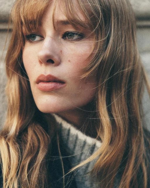 Close-up portrait of a young, fair-skinned woman with wavy blonde hair and neutral expression, wearing a grey sweater against a grey wall.
