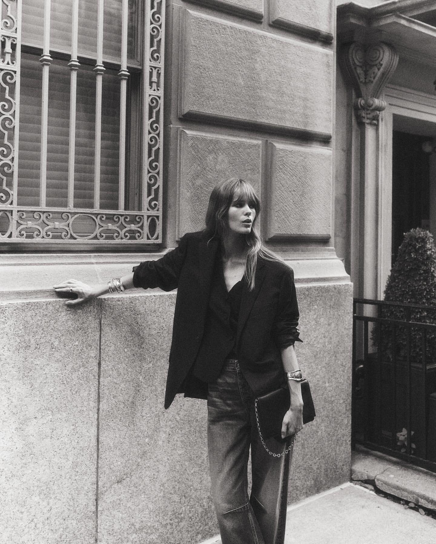 An adult woman in a black blazer, blouse, and jeans stands outside an ornate building, holding a black purse, in a black and white portrait.