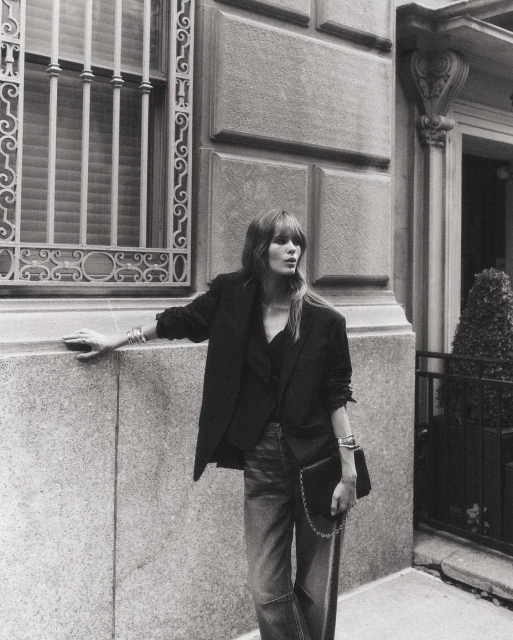 An adult woman in a black blazer, blouse, and jeans stands outside an ornate building, holding a black purse, in a black and white portrait.