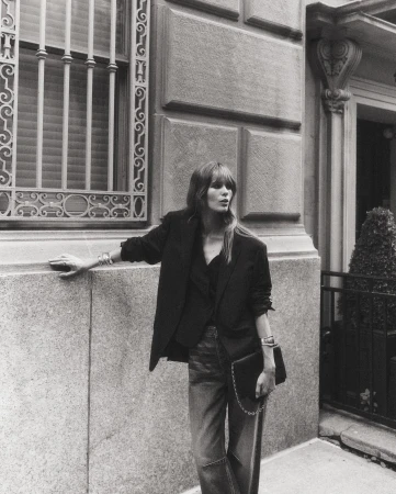 An adult woman in a black blazer, blouse, and jeans stands outside an ornate building, holding a black purse, in a black and white portrait.