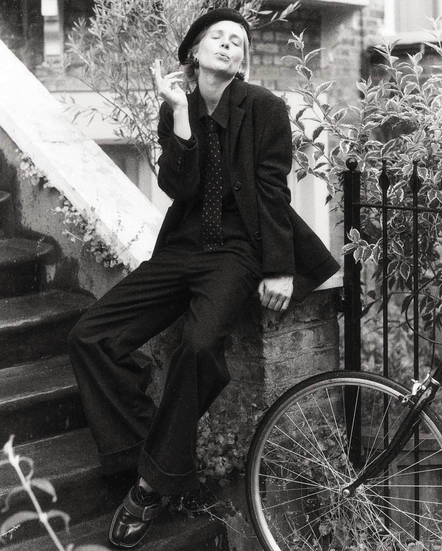 Black and white portrait of woman in suit and beret sitting on ledge, smoking cigarette, with bicycle in the foreground.