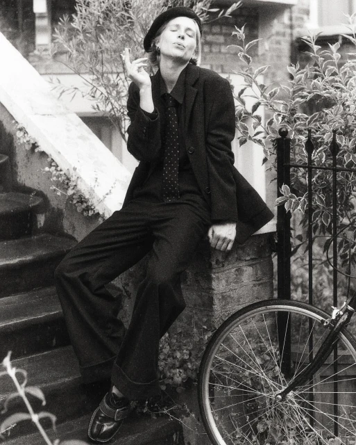 Black and white portrait of woman in suit and beret sitting on ledge, smoking cigarette, with bicycle in the foreground.