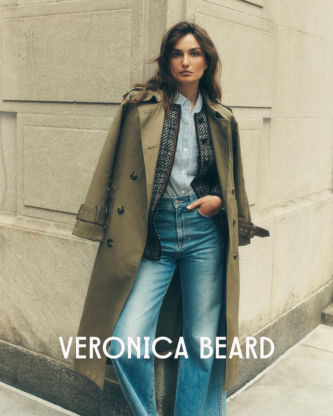 A stylish woman poses outdoors in an olive trench coat, striped shirt, jeans, and a patterned jacket with the brand VERONICA BEARD visible.