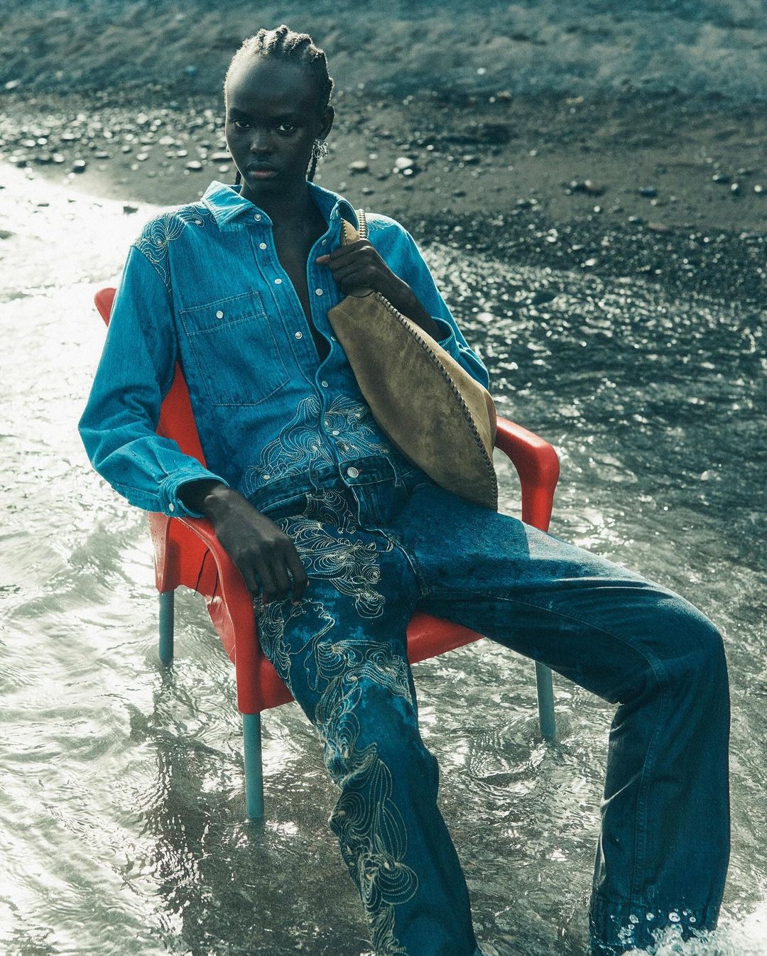 Woman in embroidered denim outfit sitting on an orange plastic chair in shallow water, a grey sandy beach in the background.