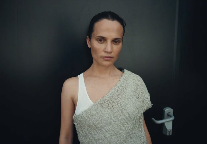 Adult female in a white tank top and grey knit shawl posing against a dark grey wall in an indoor setting, captured in a medium shot.