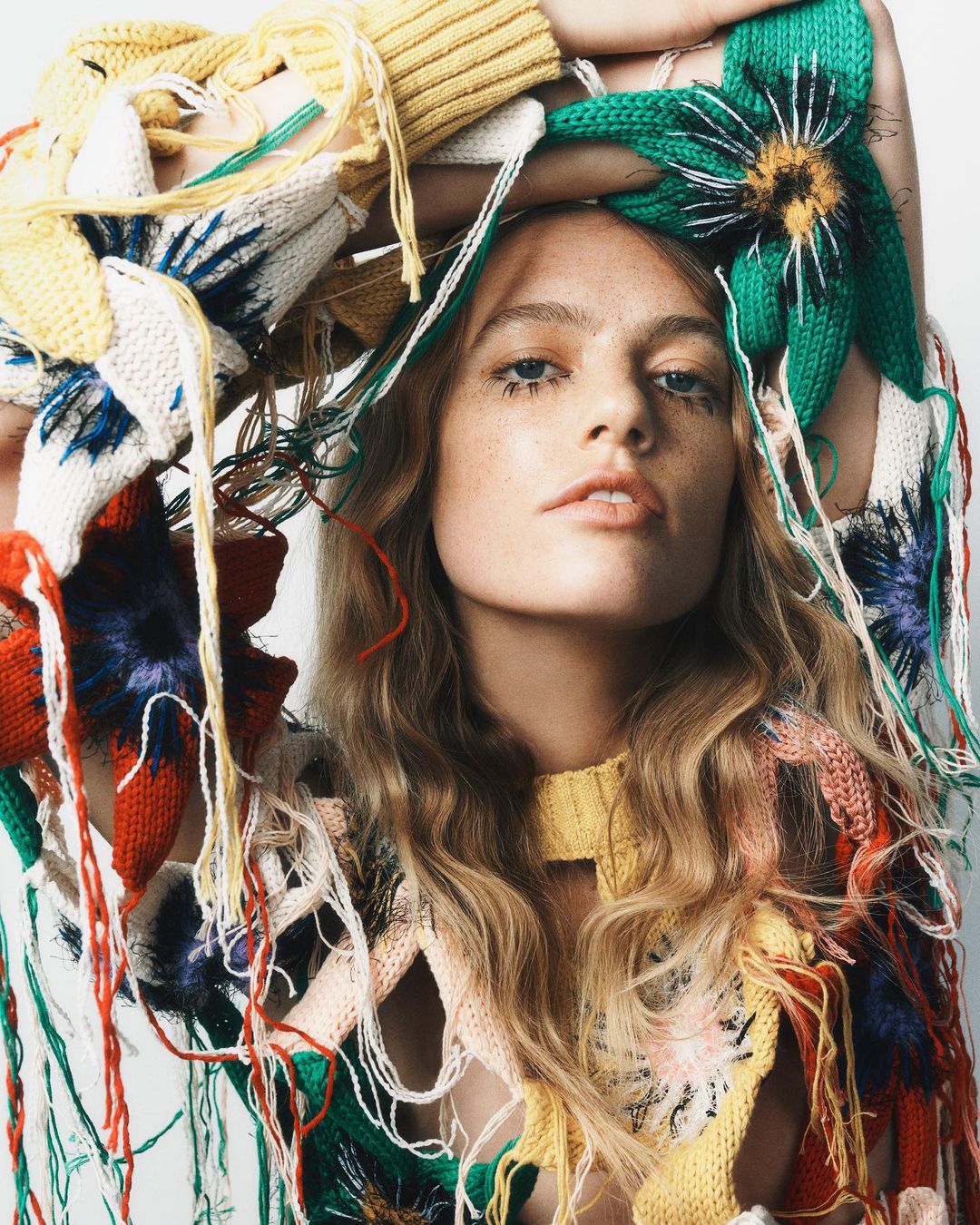 Fashionable portrait of a model wearing a colorful, floral-patterned knit top with loose strands, set against a plain, bright background.