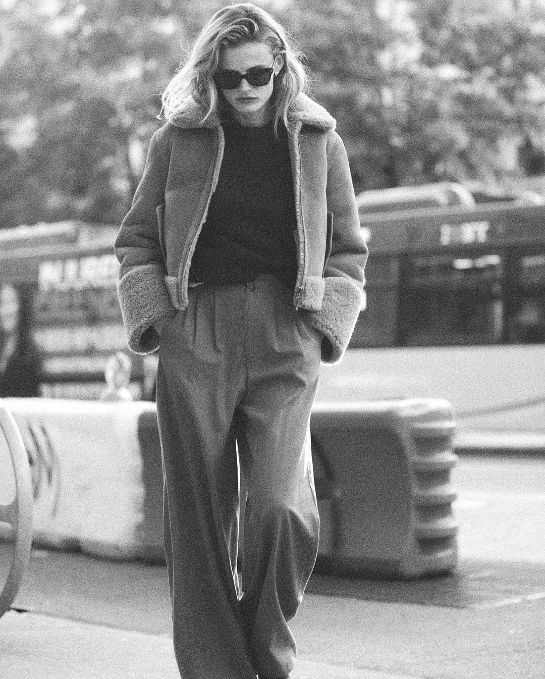 Black and white portrait of a woman walking outdoors in a shearling jacket, dark sunglasses, and wide-legged pants on a city street.