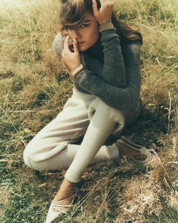 An adult female model sits in tall grass wearing a grey, long-sleeved shirt and beige leggings, styled with elegant strappy shoes.