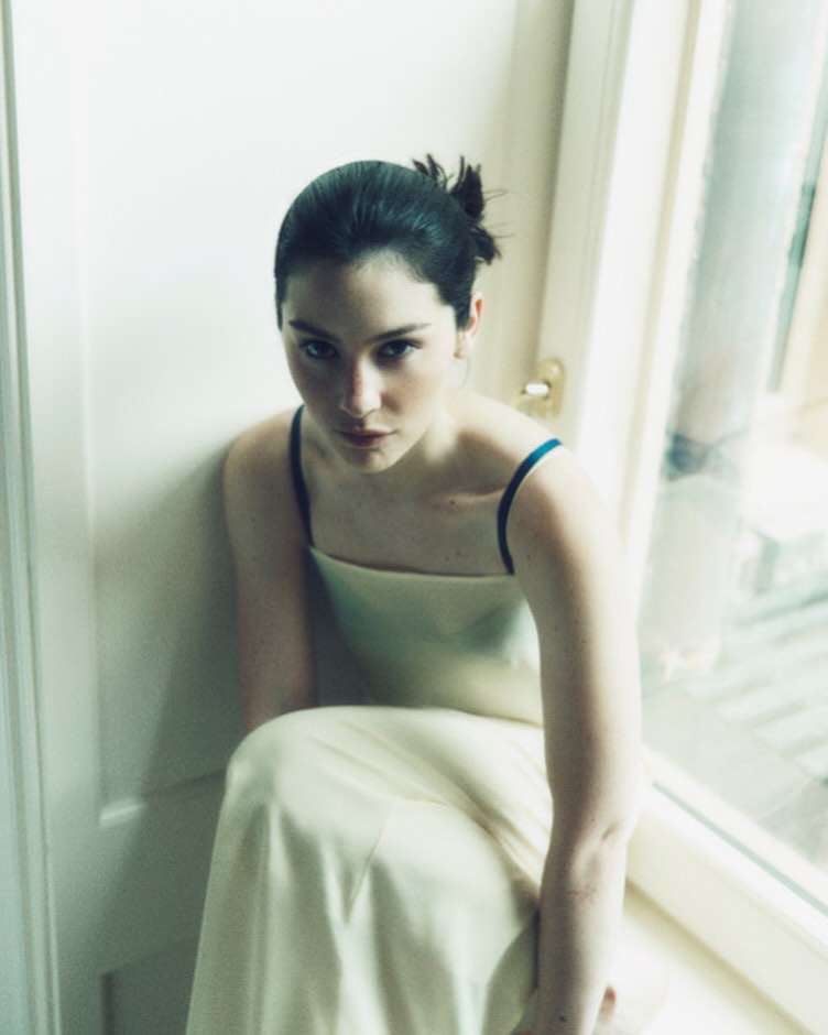 Portrait of a young woman with dark hair in a light yellow dress sitting indoors by a window with natural lighting.