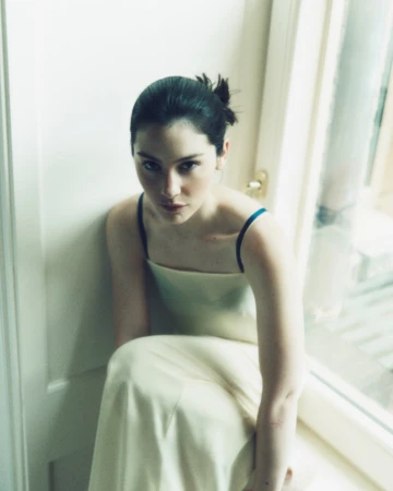 Portrait of a young woman with dark hair in a light yellow dress sitting indoors by a window with natural lighting.