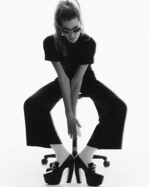 A stylish woman in a black outfit sits on a rolling chair, wearing high-heeled platform shoes, sunglasses, and socks against a white backdrop.