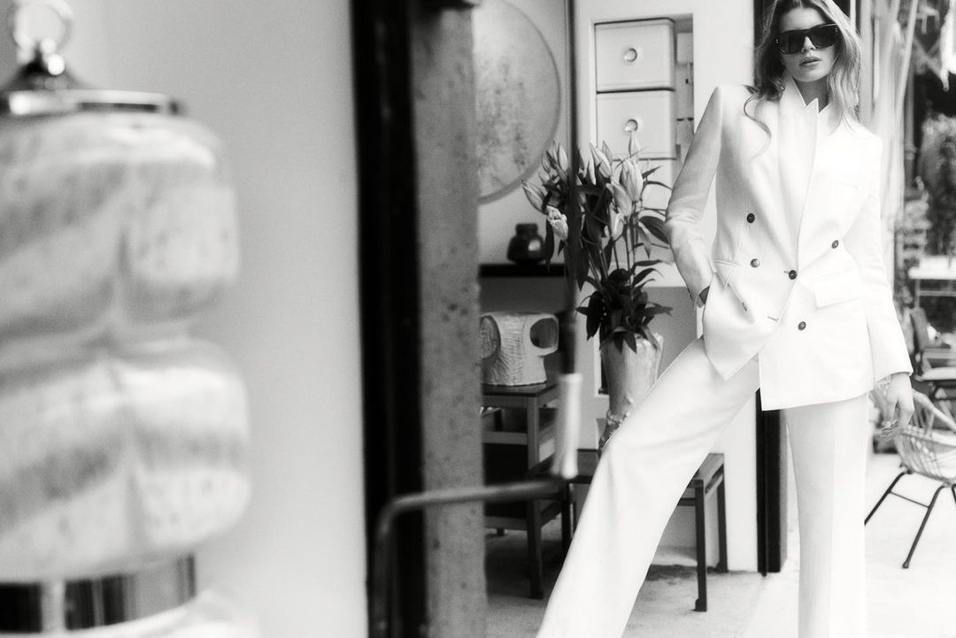 An adult woman poses in a white pantsuit and sunglasses in an indoor setting with natural light in a black and white fashion shot.