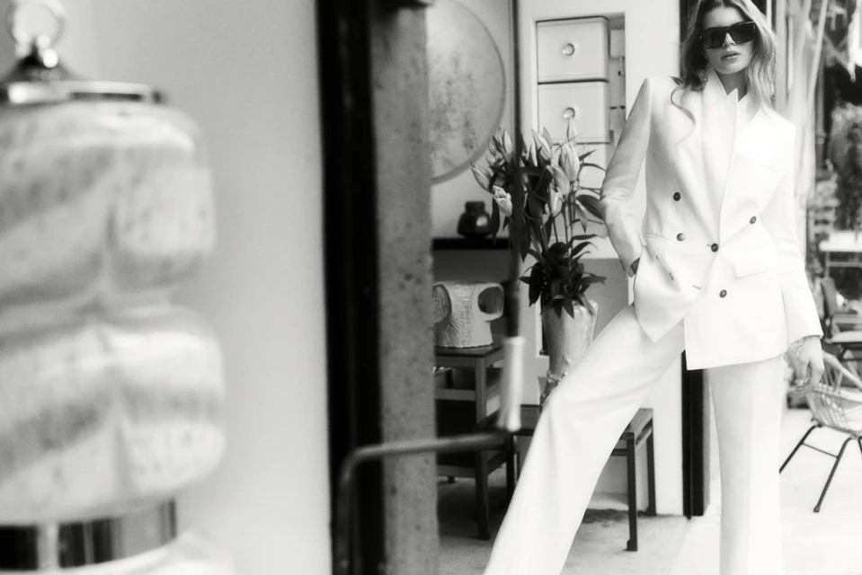 An adult woman poses in a white pantsuit and sunglasses in an indoor setting with natural light in a black and white fashion shot.