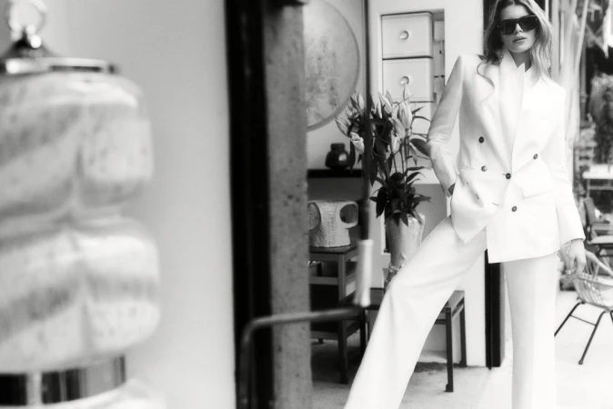 An adult woman poses in a white pantsuit and sunglasses in an indoor setting with natural light in a black and white fashion shot.