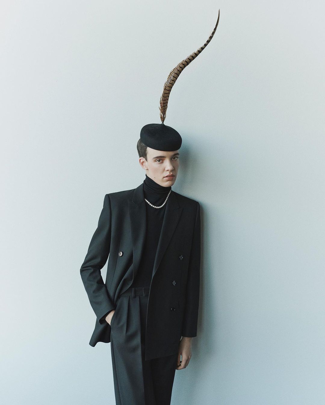 A person in a dark suit with a turtleneck and feathered hat leans against a white wall, photographed in soft, even lighting.
