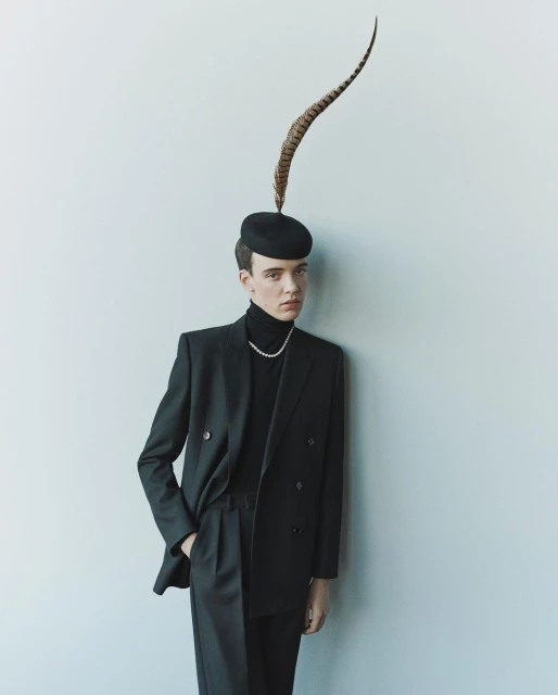 A person in a dark suit with a turtleneck and feathered hat leans against a white wall, photographed in soft, even lighting.